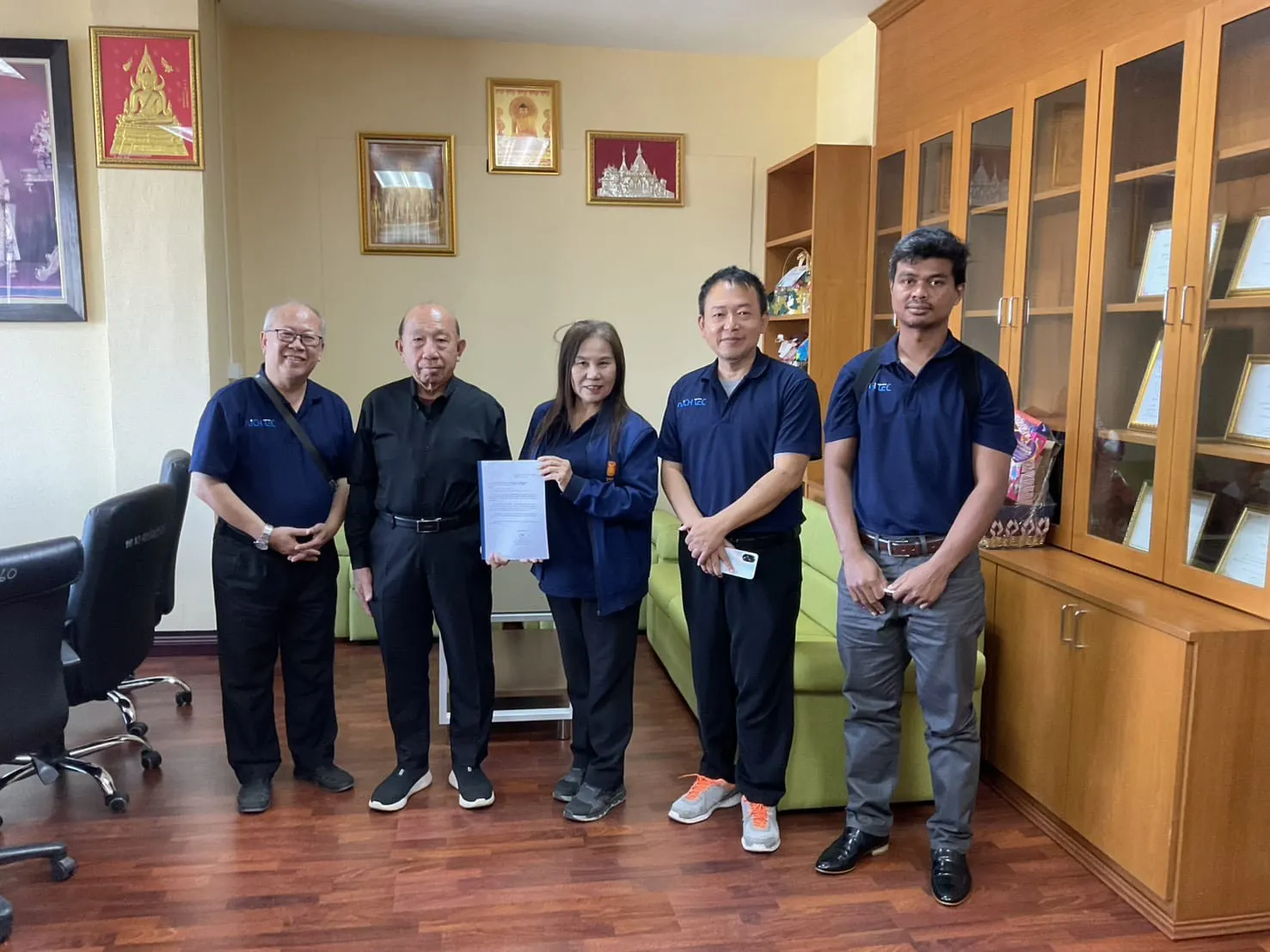 ทีม NICH TEC สามคนในเสื้อโปโลสีกรมท่า ถ่ายรูปร่วมกับเจ้าหน้าที่หน่วยงานที่ถือเอกสาร ในห้องทำงานของหน่วยงาน
